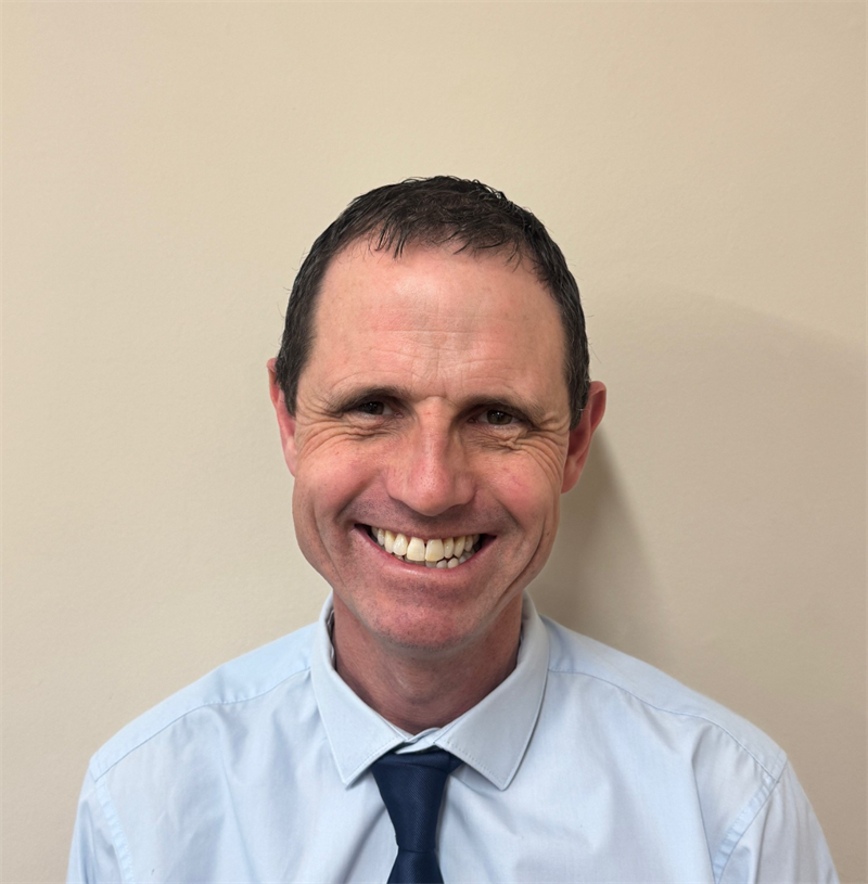 Photograph of a man with short brown hair wearing a blue shirt and navy tie. He has a friendly smile and stands against a plain, light-coloured background.