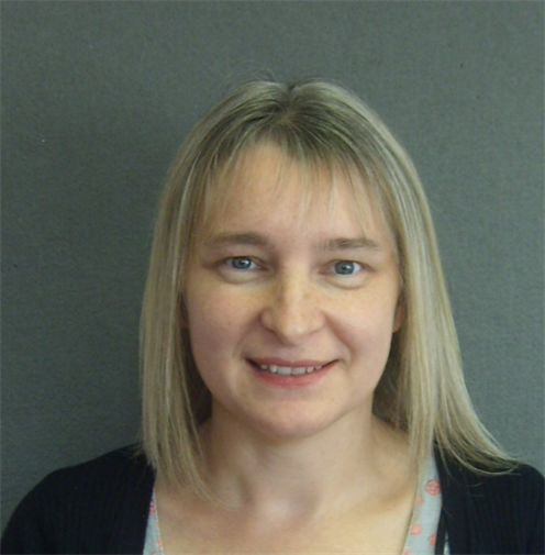 Photograph of a blonde woman with wearing a black cardigan. She has a friendly smile and stands against a plain, grey-coloured background.
