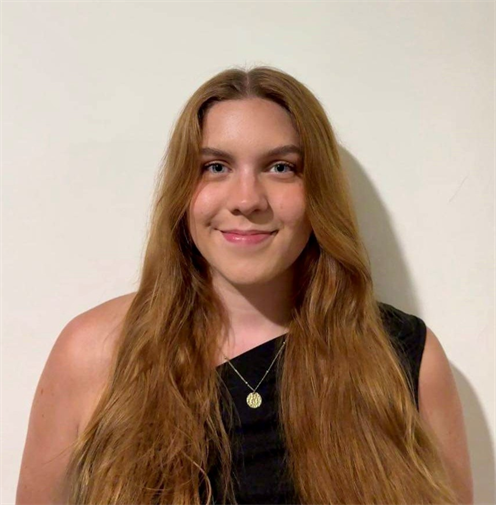 Photograph of a young woman with light brown hair in a black top. She has a friendly smile and stands against a plain, light-coloured background.