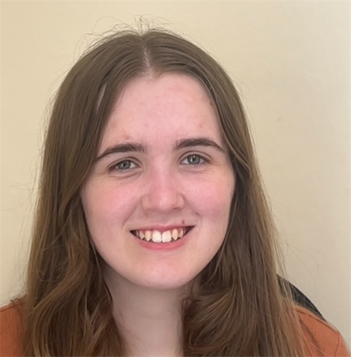 Photograph of a young woman with brown hair. She has a friendly smile and stands against a plain, light-coloured background.