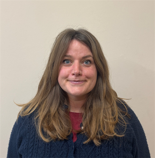Photograph of a woman with shoulder-length light brown hair wearing a blue cardigan. She has a friendly epression and stands against a plain, light-coloured background.