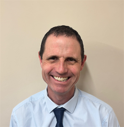 Photograph of a man with short brown hair wearing a blue shirt and navy tie. He has a friendly smile and stands against a plain, light-coloured background.