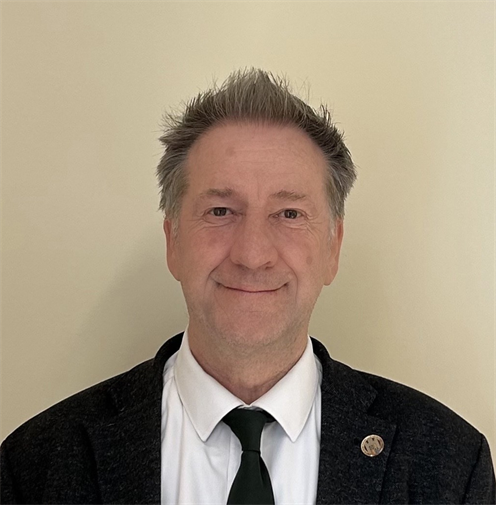 Photograph of a man with short hair, wearing a dark blazer, white shirt, and green tie. He has a neutral, friendly expression and stands against a plain, light-coloured background. 